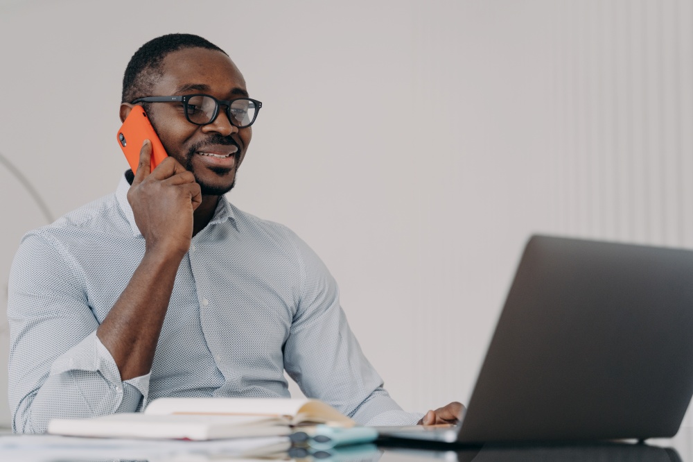 African american businessman in glasses makes pleasant business call working at laptop, holding smartphone. Smiling black male manager consulting client, talking on phone, sitting at office desk.. African american businessman in glasses makes business call, talking on phone, working at laptop