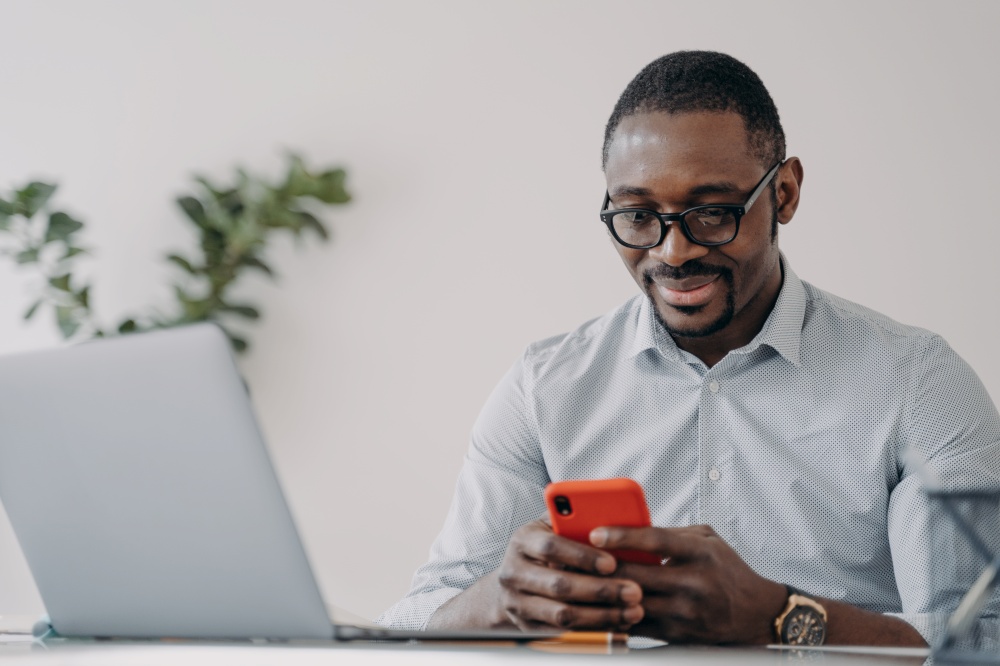 African american businessman working online using modern business apps, holding smartphone, sitting at laptop. Black businessperson checking social media job news looking at phone screen at office.. African american businessman, holding phone, uses apps for e-commerce business, working online