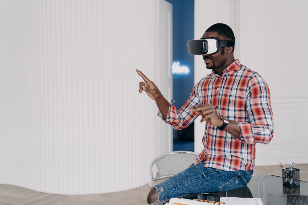 African american man in VR glasses works in augmented cyberspace, interacting with objects, sitting on office desk. Black guy uses virtual reality goggles for watching visualization of working project. African american man wearing virtual reality glasses working in augmented cyberspace in office