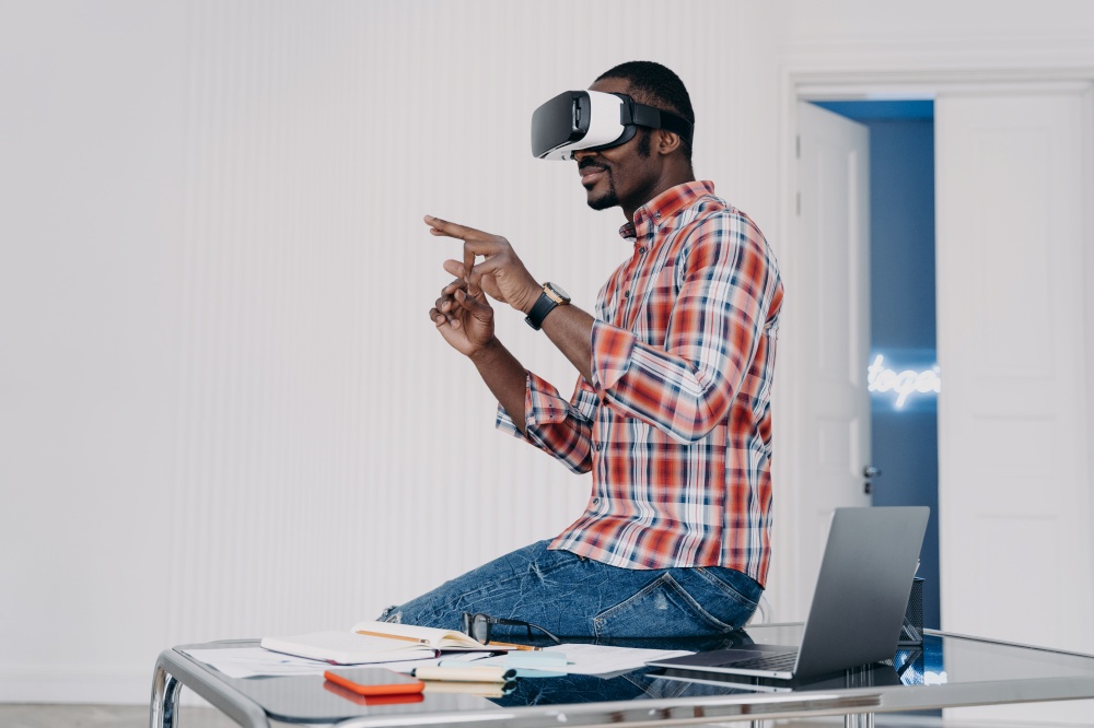 African american man in VR glasses interacts with augmented reality objects, working in office, sitting on desk. Black guy uses virtual reality goggles for job pointing in air with fingers.. African american man in VR glasses interacts with virtual reality working in office, sitting on desk