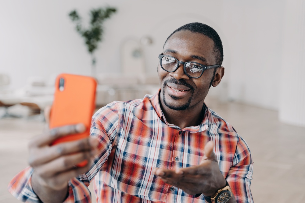 Smiling african american man wearing glasses holding phone using modern apps talking online by video call. Friendly young black guy communicating in social networks, looking at smartphone screen.. African american man in glasses holding smartphone uses modern apps talking online by video call