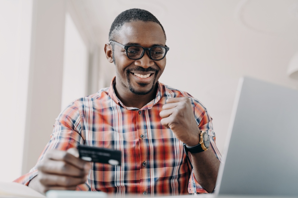 African american guy holding credit card makes yes gesture using e banking service at laptop. Black man satisfied by successful financial transaction with cashback. Online banking, e commerce.. African american man using online banking service at laptop, holding credit card makes yes gesture