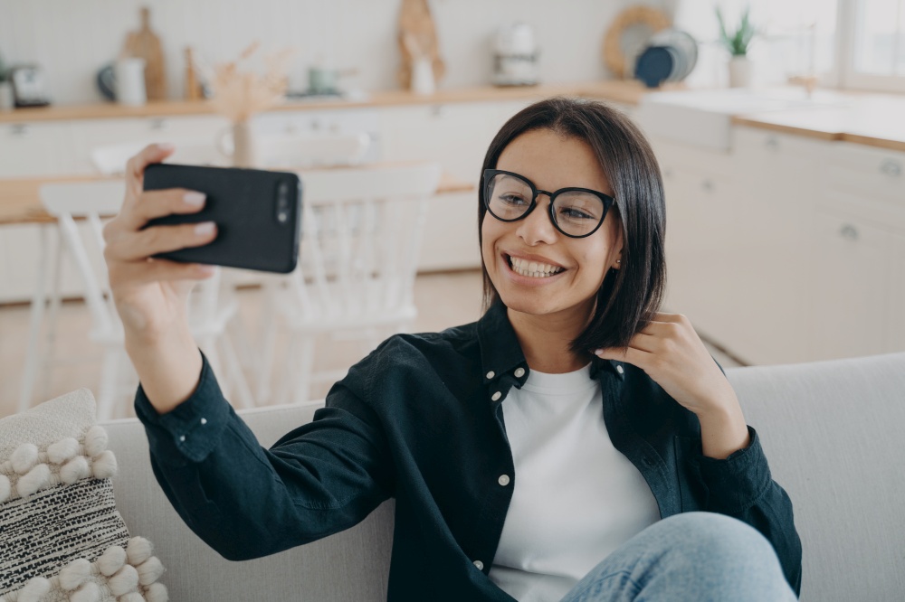 Smiling girl blogger holding smartphone, talks by video call, takes selfie photos, sitting on couch at home. Happy young woman wearing glasses chatting online with followers in social networks.. Smiling girl blogger holding smartphone, talks by video call, takes selfie photos, sitting on couch