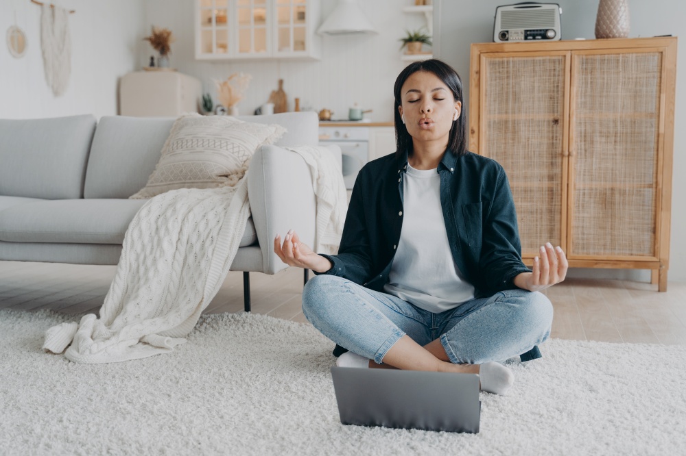 Female practices yoga at laptop, breathing deep sitting on floor in living room. Calm woman meditates after working day, listening online meditation at home. Stress relief, healthy lifestyle.. Female practices yoga at laptop, breathing deep sitting on floor at home. Stress relief, meditation