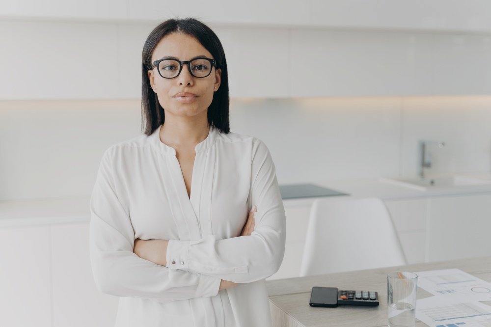 Professional manager woman wearing glasses standing with her arms crossed looking at camera. Confident businesswoman, corporate leader, boss, principal posing in office on workplace.. Businesswoman, corporate leader, director wearing glasses standing with her arms crossed in office