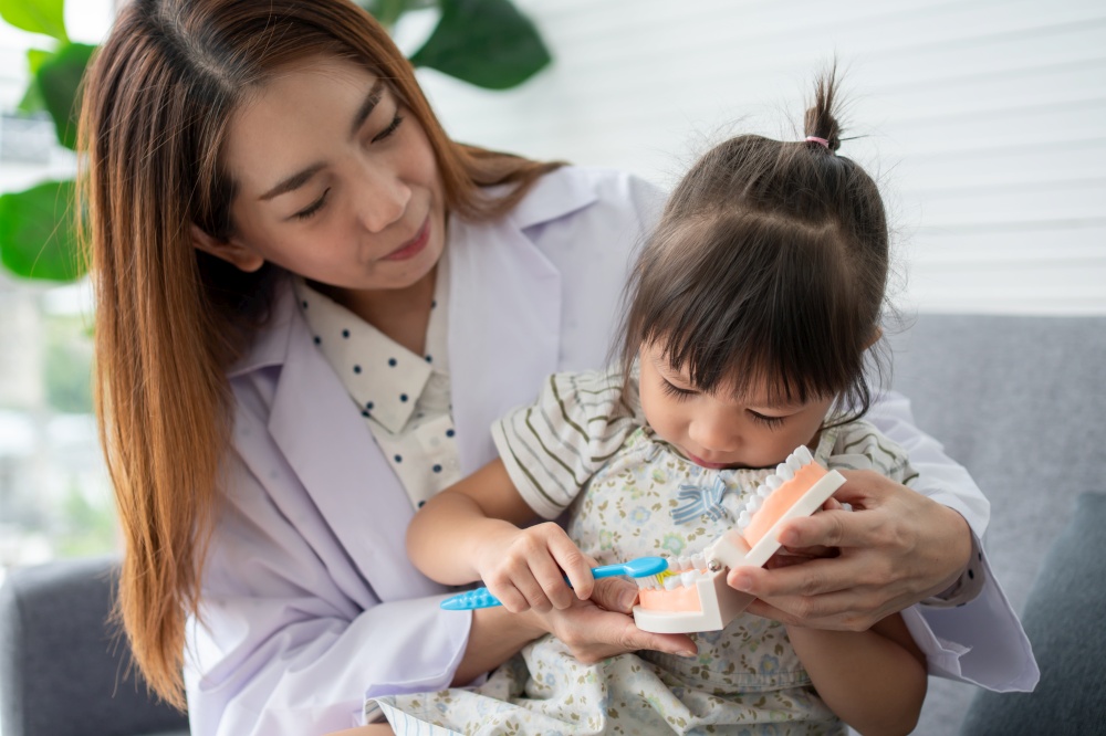 Asian female dentist teaching cute little girl brushing teeth with toothbrush and stomatologist telling girl child about oral hygiene in dental clinic, Education and prevention cavities concept.