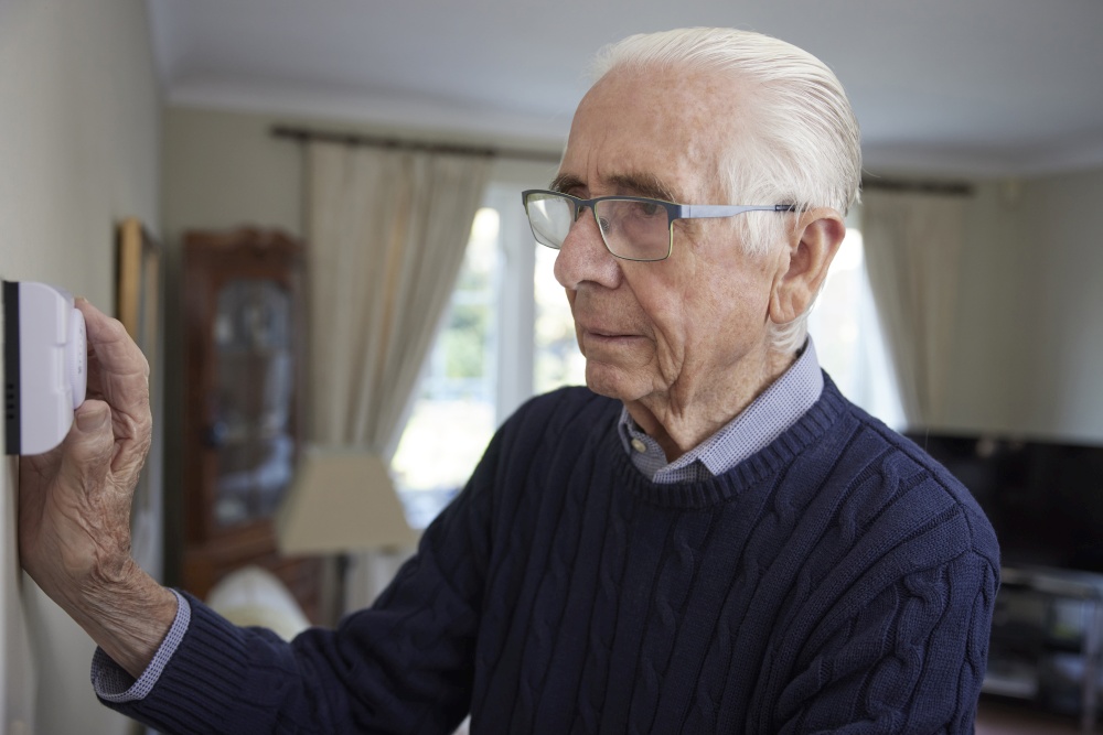 Worried Senior Man Turning Down Central Heating Thermostat At Home In Energy Crisis
