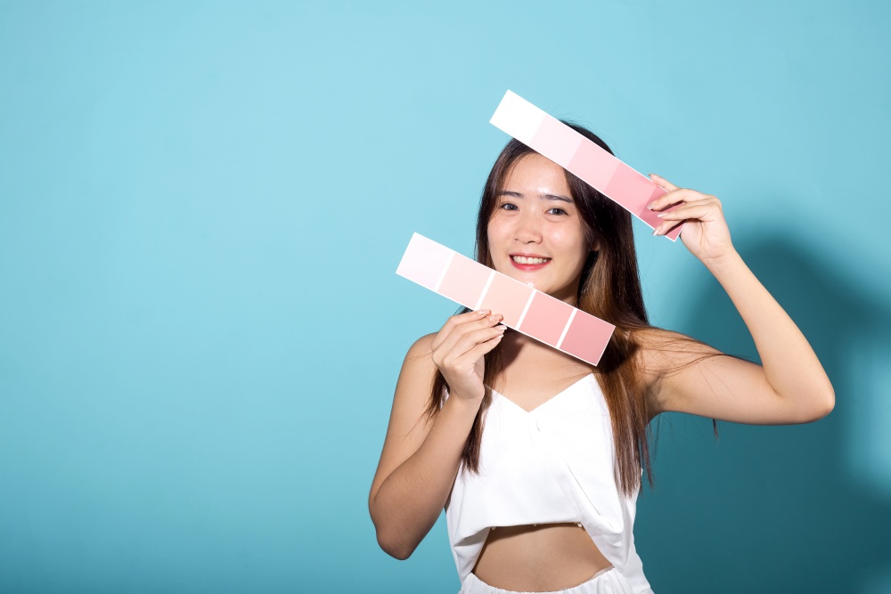 Asian beautiful young woman holding skin color scale paper studio shot isolated on blue background, Portrait of happy female hold color card under sun protection, skin beauty face