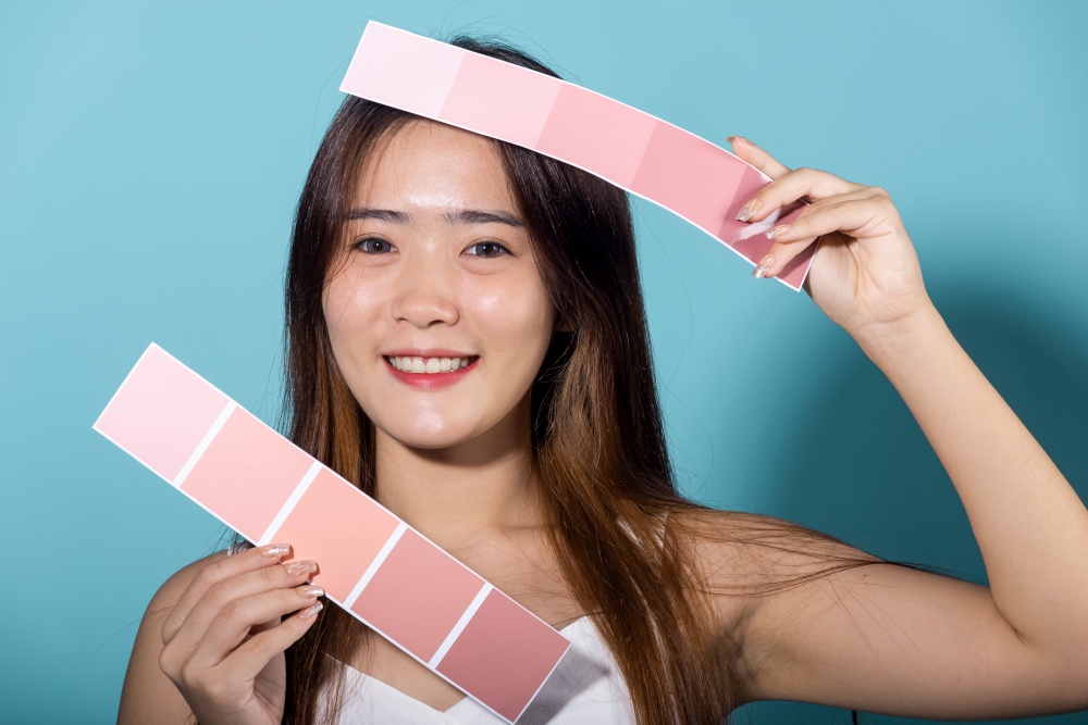 Portrait of happy female hold color card under sun protection, skin beauty face, Asian beautiful young woman smiling and holding skin color scale paper studio shot isolated on blue background
