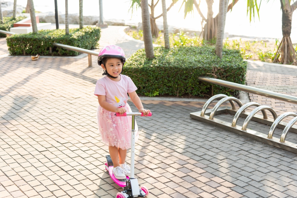 Child riding scooter. Happy Asian little kid girl wear safe helmet playing pink kick board on road in park outdoors on summer day, Active children games outside, Kids sport healthy lifestyle concept