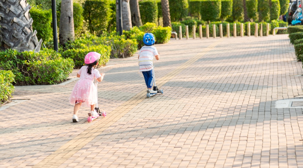 Child riding scooter. Back Asian little kid boy and girl wear safe helmet play kick board on road in park outdoors on summer day, Active children games outside, Kids sport healthy lifestyle concept