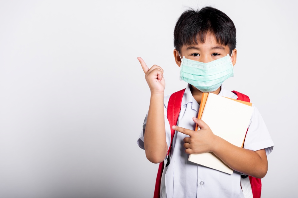 Asian student kid boy wear student thai uniform and protect face mask ready to go to school pointing finger to space in studio shot isolated on white background, preschool, new normal back to school