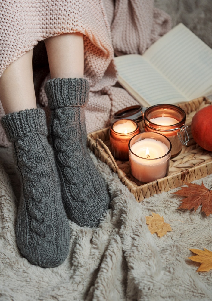 The girl is covered with a blanket in knitted socks. Cozy autumn concept. Pumpkins and candles on a tray near the girl.