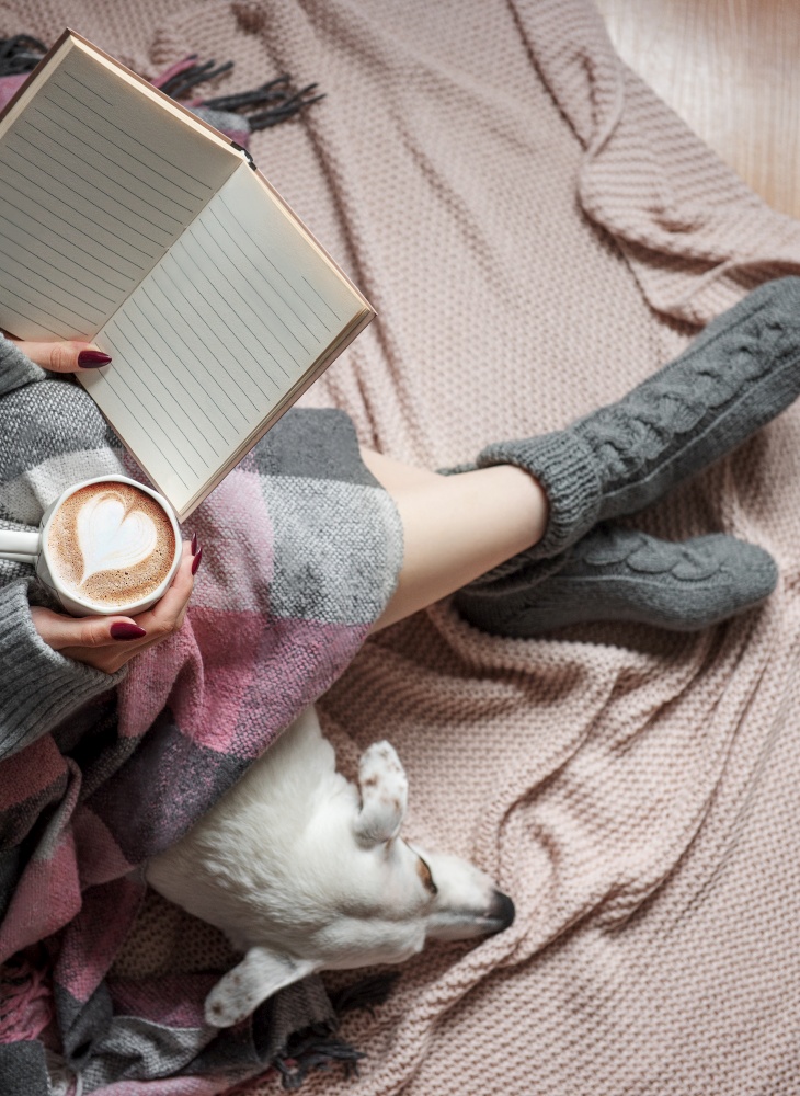 Cozy home, woman covered with warm blanket, drinks coffee,  sleeping dog next to woman. Relax, carefree, comfort lifestyle.