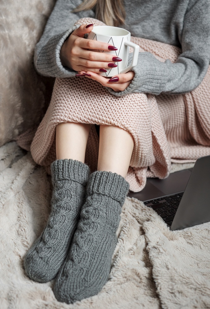 Cozy home, woman covered with warm blanket, drinks coffee and works on a laptop. Relax, carefree, comfort lifestyle.