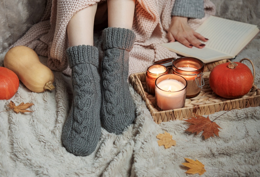 The girl is covered with a blanket in knitted socks. Cozy autumn concept. Pumpkins and candles on a tray near the girl.