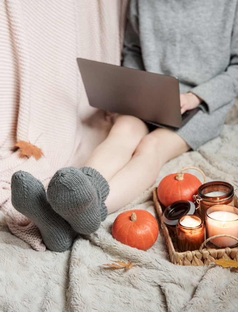 A girl in a sweater and knitted socks sits on a blanket and works on a laptop. Near her is a tray with all things and pumpkins. Cozy autumn concept.