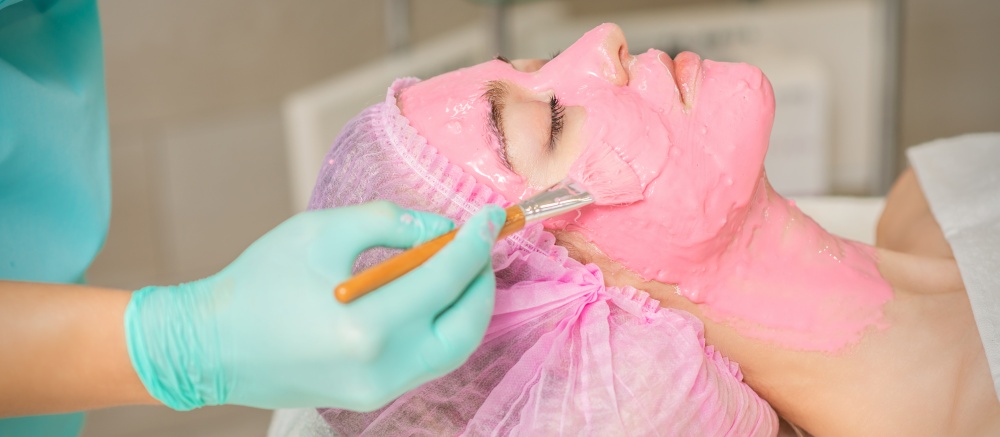 Hand of cosmetologist applying pink alginic mask to face of young woman in beauty salon. Cosmetologist applying pink alginic mask