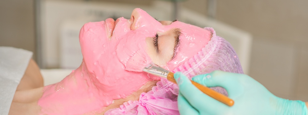 Hand of cosmetologist applying pink alginic mask to face of young woman in beauty salon. Cosmetologist applying pink alginic mask