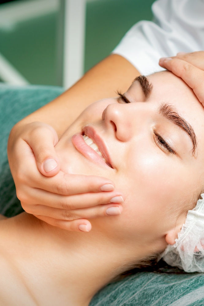 Young woman getting a facial massage with closed eyes by a beautician in a beauty salon. Young woman getting facial massage