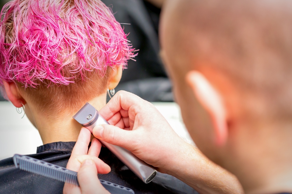 Hairdresser shaving nape and neck with electric trimmer of a young caucasian woman with short pink hair in a beauty salon. Hairdresser shaving nape and neck with electric trimmer of a young caucasian woman with short pink hair in a beauty salon.