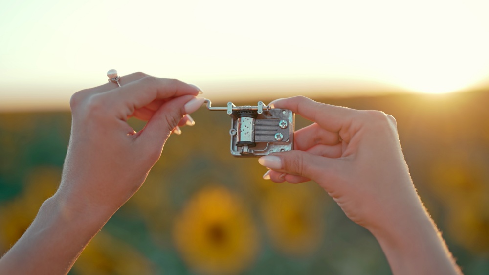 Female hands rotating gears of old music box mechanism. Lady turning the lever of retro small metallic carillon. Woman in sunflowers field listening to music which playing. High quality photo. Female hands rotating gears of old music box mechanism. Lady turning the lever of retro small metallic carillon. Woman in sunflowers field listening to music which playing.