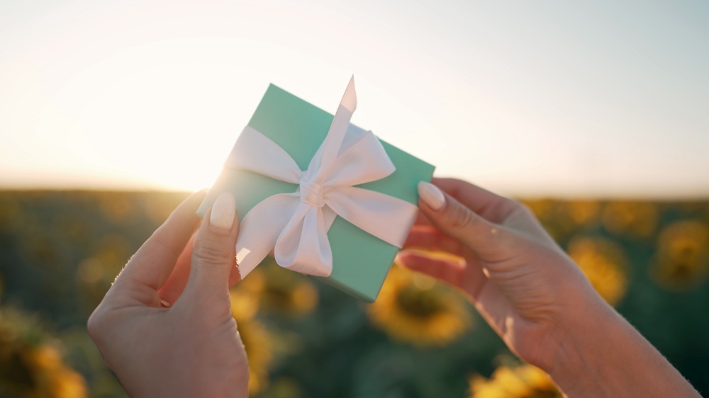 Woman holding blue gift box with bow on sunflowers field background with sun flares. Present, bonus, offer concept. Only hands. High quality photo. Woman holding blue gift box with bow on sunflowers field background with sun flares. Present, bonus, offer concept. Only hands.