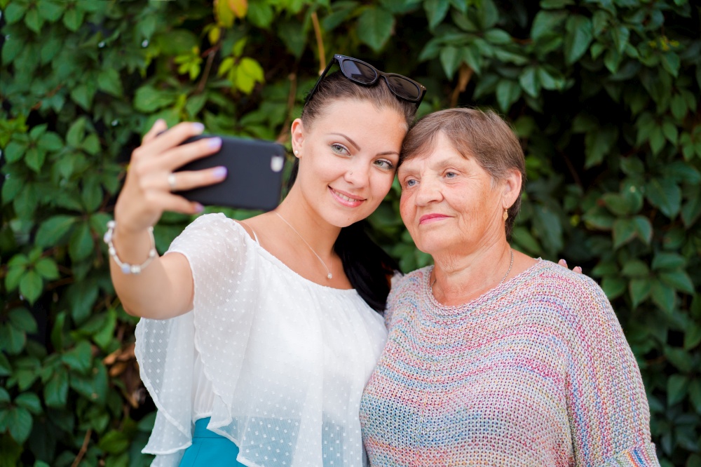 Pretty old granny and her granddaughter doing selfie outdoor, fooling around, looking at camera and laughing to smartphone camera. Technology, memory, family concept. Pretty old granny and her granddaughter doing selfie outdoor, fooling around, looking at camera and laughing to smartphone camera. Technology, memory, family concept.