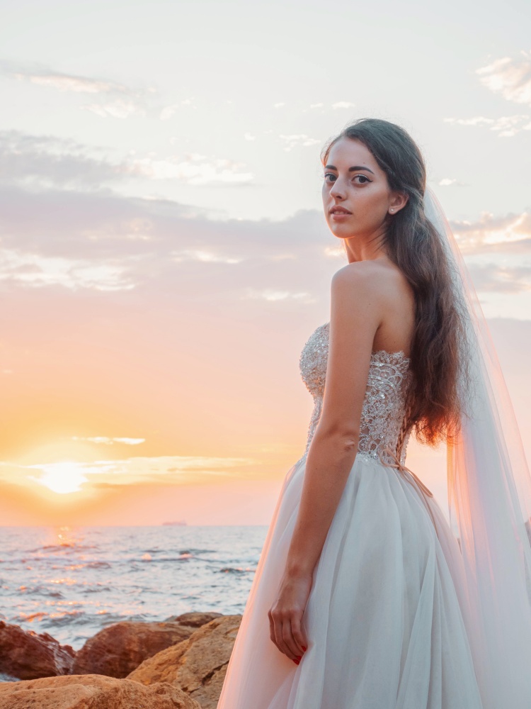 Portrait of girl in wedding luxury dress watching sunrise on sea shore. Bride on a rocks. Beautiful waves near to her. Woman enjoying happy moments with nature.