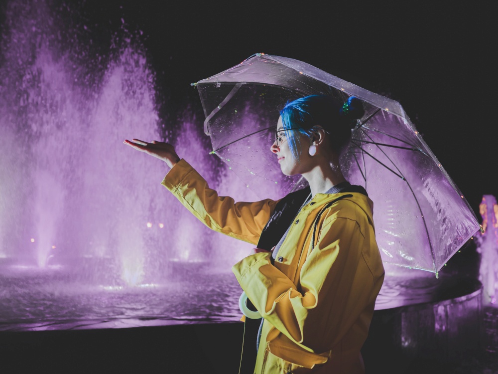 Young pretty girl with blue dyed hair in yellow raincoat and with transparent umbrella stands near fountain. Night neon illumination of city. Portrait of stylish hipster with glasses.
