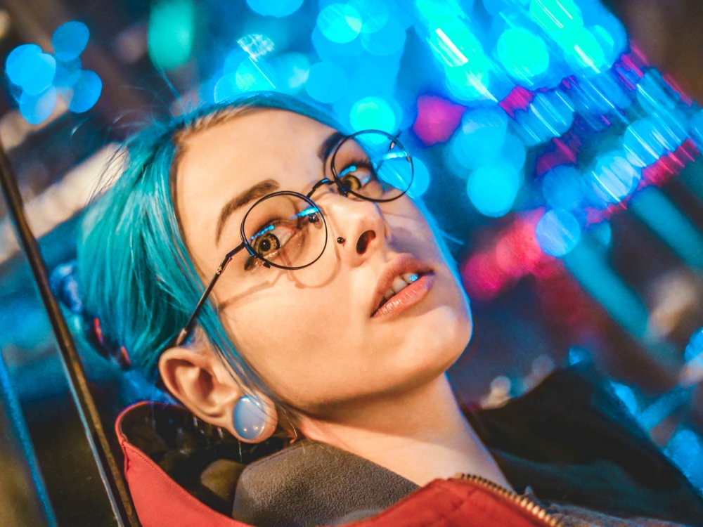 Portrait of young pretty girl with unusual hairstyle lying on glass wall which glowing colorful neon lights. City at night. Dyed blue hair in braids. Happy hipster teenager in glasses.