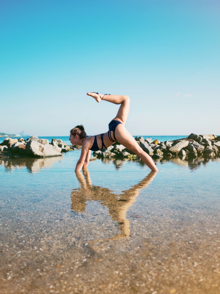 Young caucasian woman in swimsuit practicing yoga in sea water, lake or river. Beautiful reflection. Complex asanas, balance. Fitness, sport, yoga and healthy lifestyle concept.