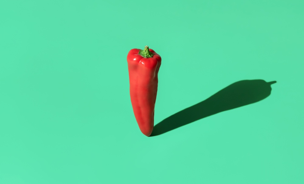 Single long red pepper minimalist on a green-colored table. Raw sweet pepper standing on a colorful background with a long strong shadow