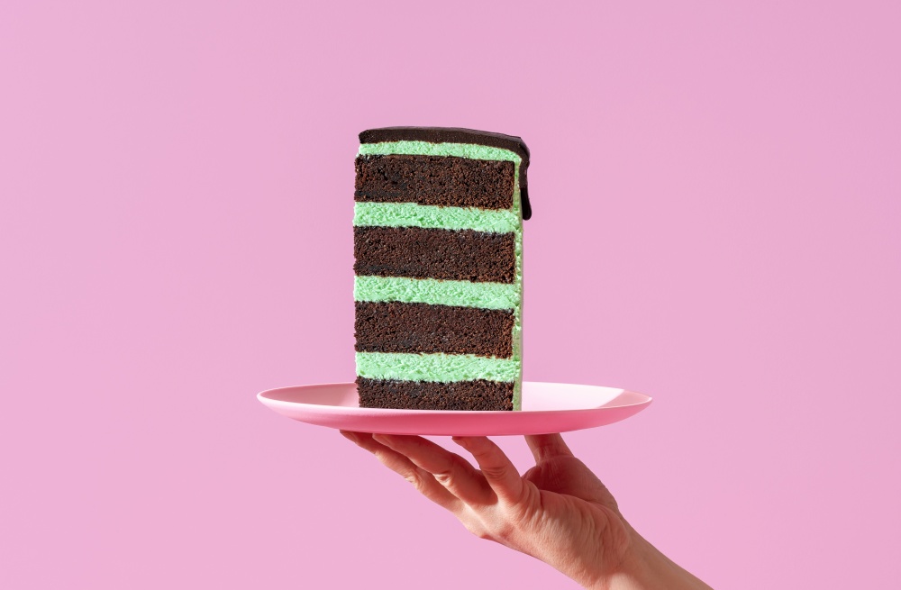 Woman&rsquo;s hand holding a plate with a slice of peppermint cake against a purple background. Homemade layer cake with mint-flavored cream and chocolate topping