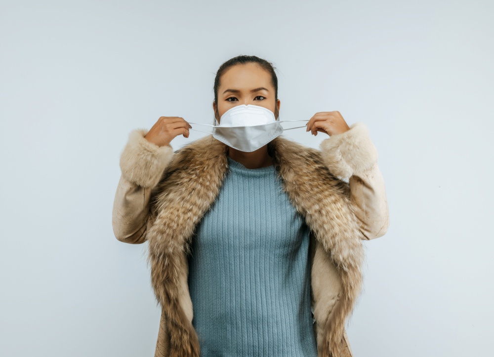 Portrait of Asian woman putting on protective mask on face for protection against coronavirus, wearing a fur coat standing over background in studio with copy space.
