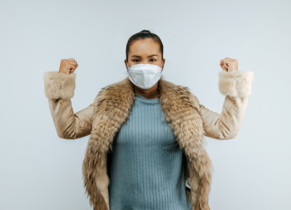 Portrait of Asian woman keeps hands clenched in fists, showing powerful, putting on protective mask on face for protection against coronavirus, wearing a fur coat, isolated over background.