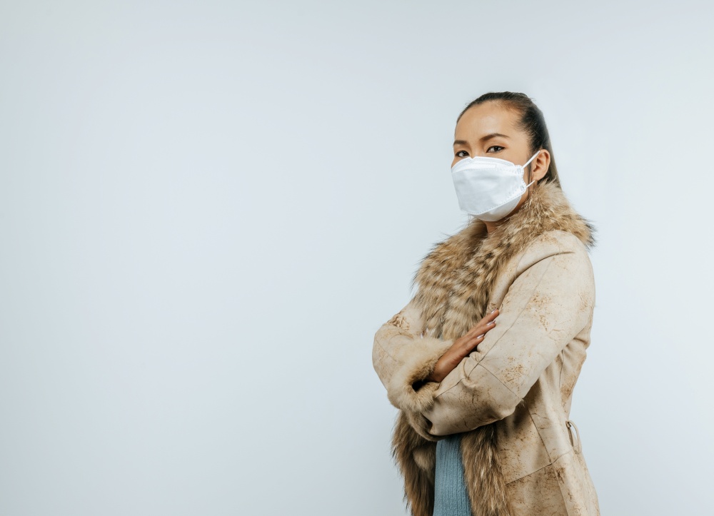 Portrait of Asian woman staring at the camera and crossed arms, putting on protective mask on face for protection against coronavirus, wearing a fur coat, isolated over background.