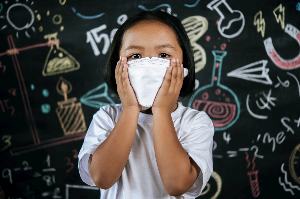 Portrait of a little girl wearing eyeglasses and facemask at school to avoid the spread of coronavirus, new normal with schoolchild,  blure blackboard in background