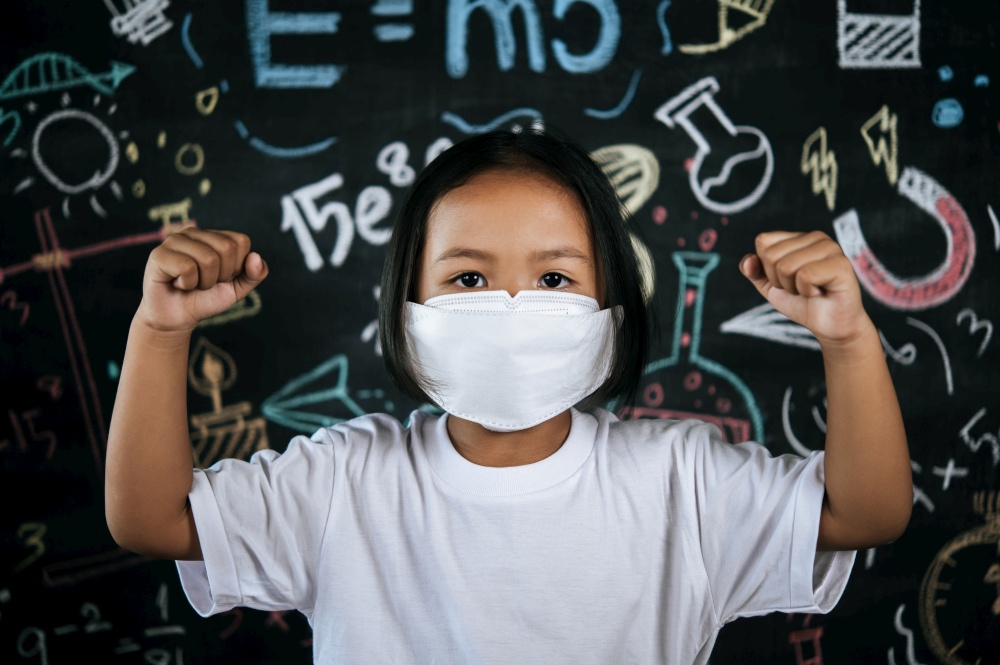 Portrait of a little girl wearing eyeglasses and facemask at school to avoid the spread of coronavirus, new normal with schoolchild,  blure blackboard in background