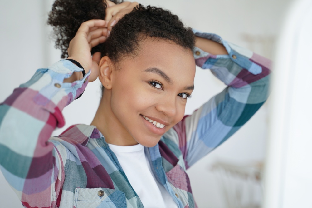 Smiling young mixed race girl holding healthy strong afro hairs, doing hairstyle, happy satisfied beautiful teen lady looking in mirror makes ponytail. Natural beauty, self care routine concept.. Smiling young mixed race girl holding healthy afro hairs, doing hairstyle. Natural beauty, self care