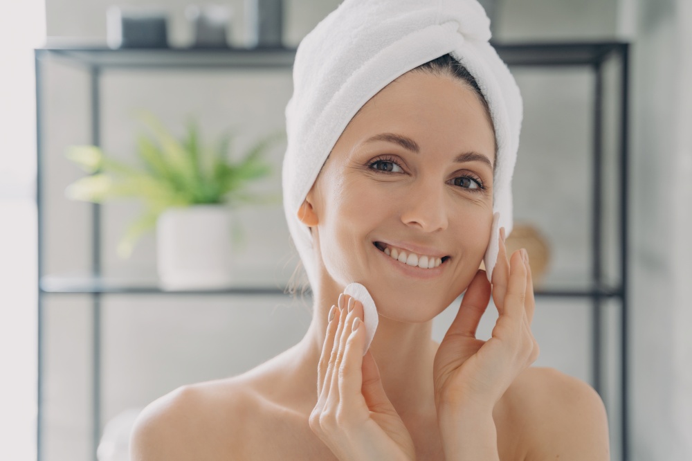 Hispanic woman in towel on head cleansing facial skin with cotton pads after shower. Smiling female enjoy healthy clean skin, remove makeup using tonic, micellar water in bathroom. Skincare concept.. Hispanic woman cleansing facial skin with cotton pads after shower removing makeup. Skincare routine