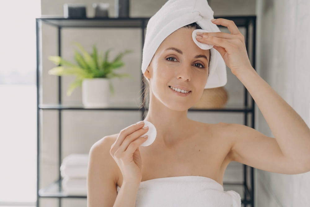 Latina woman moisturizing, cleansing facial skin, using cotton pads and micellar water or milk, removing makeup after the shower in bathroom. Self care, personal hygiene, skincare daily routine.. Woman moisturizing, cleansing facial skin, using cotton pads in bathroom. Personal hygiene, skincare