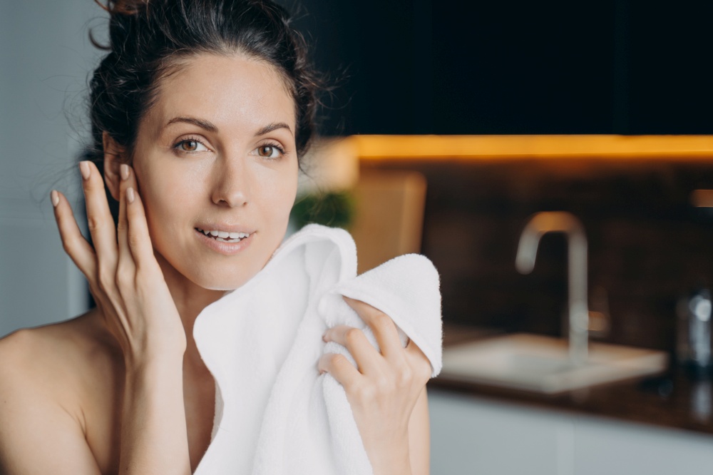 Beautiful woman wiping face with soft white towel after washing at home. Spanish female enjoying perfect healthy smooth skin after skincare beauty treatment. Hygiene, self care concept.. Beautiful woman enjoying healthy smooth skin after washing face, holding towel. Skincare at home