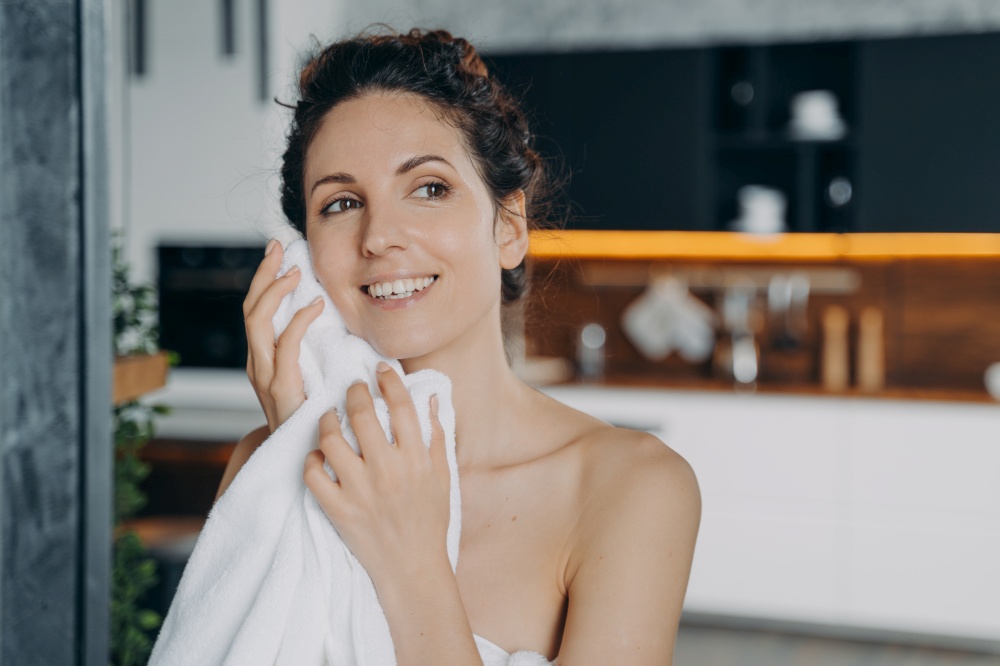 Young hispanic woman wiping face with towel after washing at home. Pretty smiling female holding towel cleansing, drying facial skin with napkin after the shower in the morning. Daily skincare routine. Woman wiping facial skin with towel after washing in the morning at home. Daily skincare routine