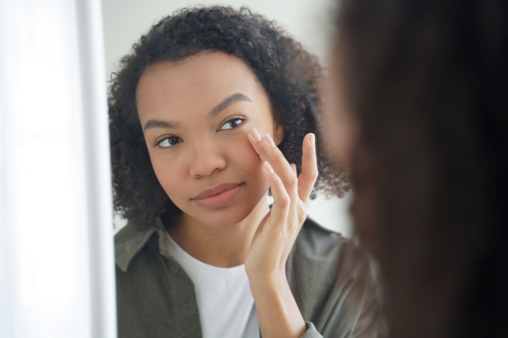 Mixed race young girl looking at mirror, touches delicate under eye skin after applying moisturizing treatment cosmetics. Pretty biracial lady apply daily facial cream. Natural beauty and skincare.. Mixed race girl touches delicate under eye skin, applying facial cream. Natural beauty, skincare