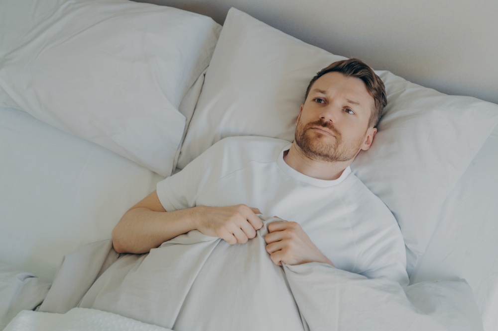 View from above of lonely upset young bearded man lying in bed with open eyes and can&rsquo;t sleep, feeling unhappy and tired, squeezes blanket with hands and being stressed of insomnia. Sleeping problems. Lonely upset young bearded man lying in bed with open eyes