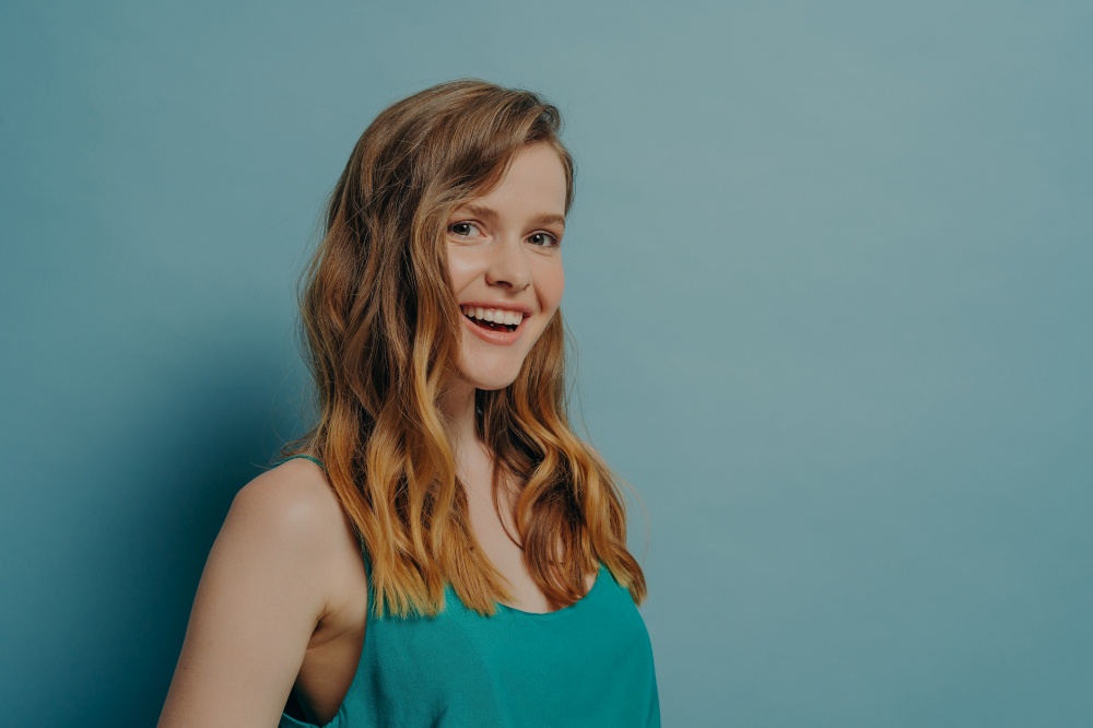 Portrait of lovely attractive millenial girl with wavy perfectly styled hair standing sideways looking at camera with joyful radiant smile isolated over pastel blue studio wall background. Positive millenial girl with wavy hair stands sideways and looks at camera with joyful radiant smile