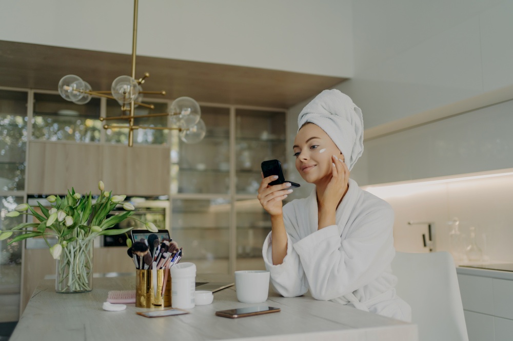 Young attractive woman in bathrobe with hair wrapped in towel applying cosmetic cream on face and looking in compact mirror, doing cosmetic procedures for skin while sitting in modern kitchen at home. Happy healthy woman in bathrobe taking care of her skin after taking shower or bath at home
