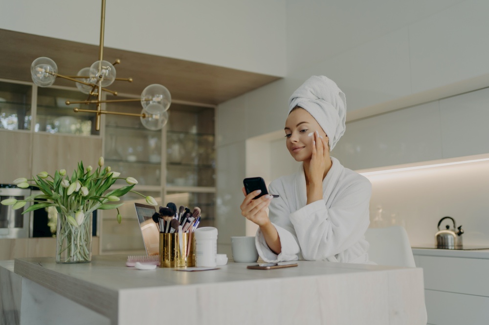 Beauty and facial care concept. Young pretty woman in white bathrobe applying cosmetic product on face and looking in compact mirror, taking care of her skin while sitting in modern kitchen at home. Happy healthy woman in bathrobe doing skincare daily routine after taking shower or bath at home