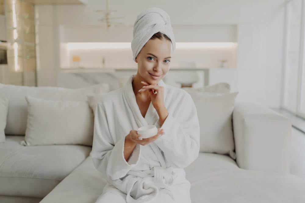 Skincare and beauty concept. Portrait of young attractive woman in bathrobe and towel on head holding cream jar and smiling at camera while sitting on sofa in living room doing cosmetic procedures. Young happy woman holding cosmetic face cream and smiling at camera while relaxing on sofa at home